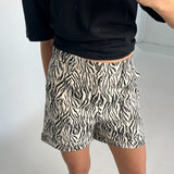 En person iført en sort trøje fotograferer sig selv iført zebrastribede shorts i et minimalistisk, hvidt rum.