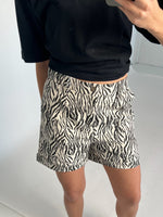 En person iført en sort trøje fotograferer sig selv iført zebrastribede shorts i et minimalistisk, hvidt rum.