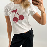 En person iført hvid t-shirt med rødt kirsebærmotiv tager en selfie i et spejl. Personen har sort bukser, rød neglelak, gyldent armbånd og holder en brun telefon.