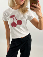 En person iført hvid t-shirt med rødt kirsebærmotiv tager en selfie i et spejl. Personen har sort bukser, rød neglelak, gyldent armbånd og holder en brun telefon.