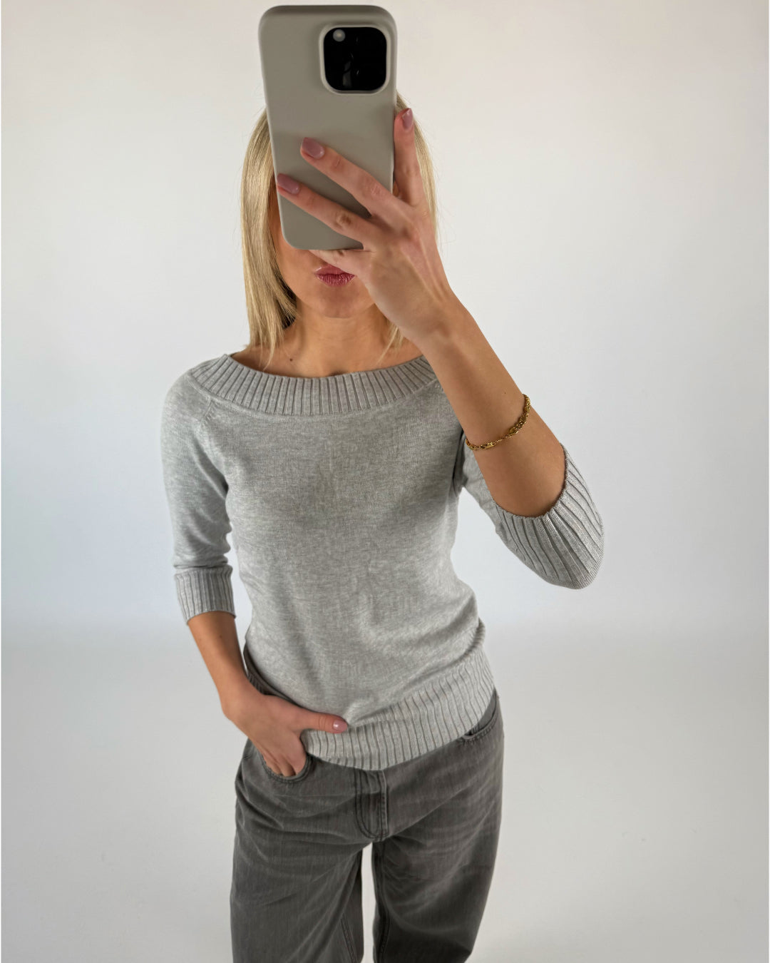 En person tager en selfie foran en hvid baggrund, iført en grå sweater og jeans, med hånden i lommen og en mobiltelefon foran ansigtet.