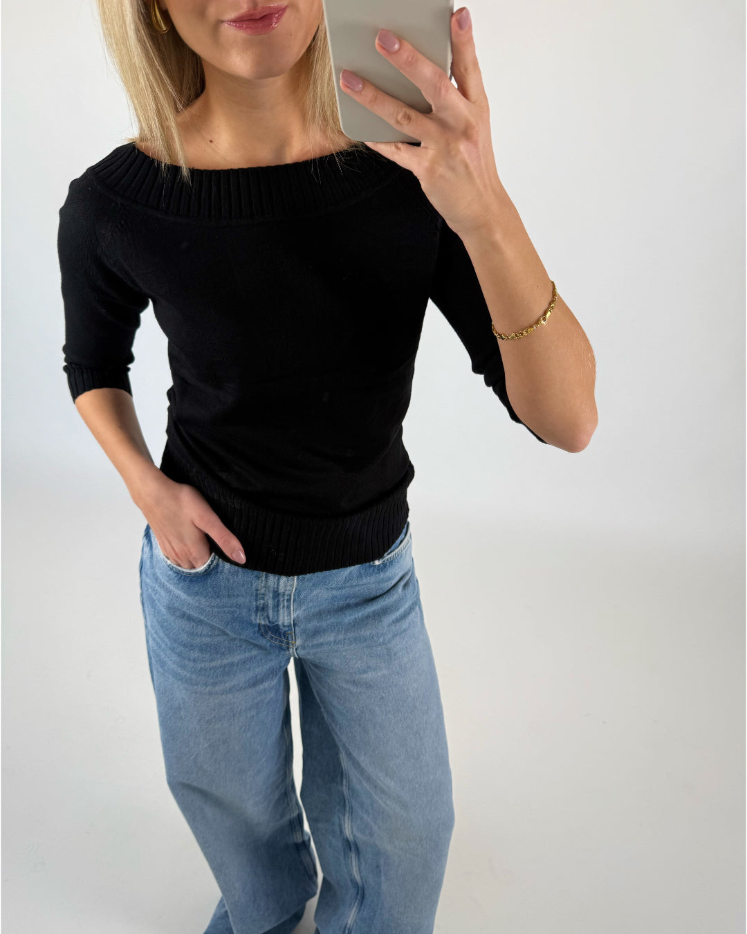 En kvinde iført sort trøje og lyse jeans tager et spejlbillede med sin smartphone i et lyst rum. Hun har en hånd i lommen og bærer et armbånd.