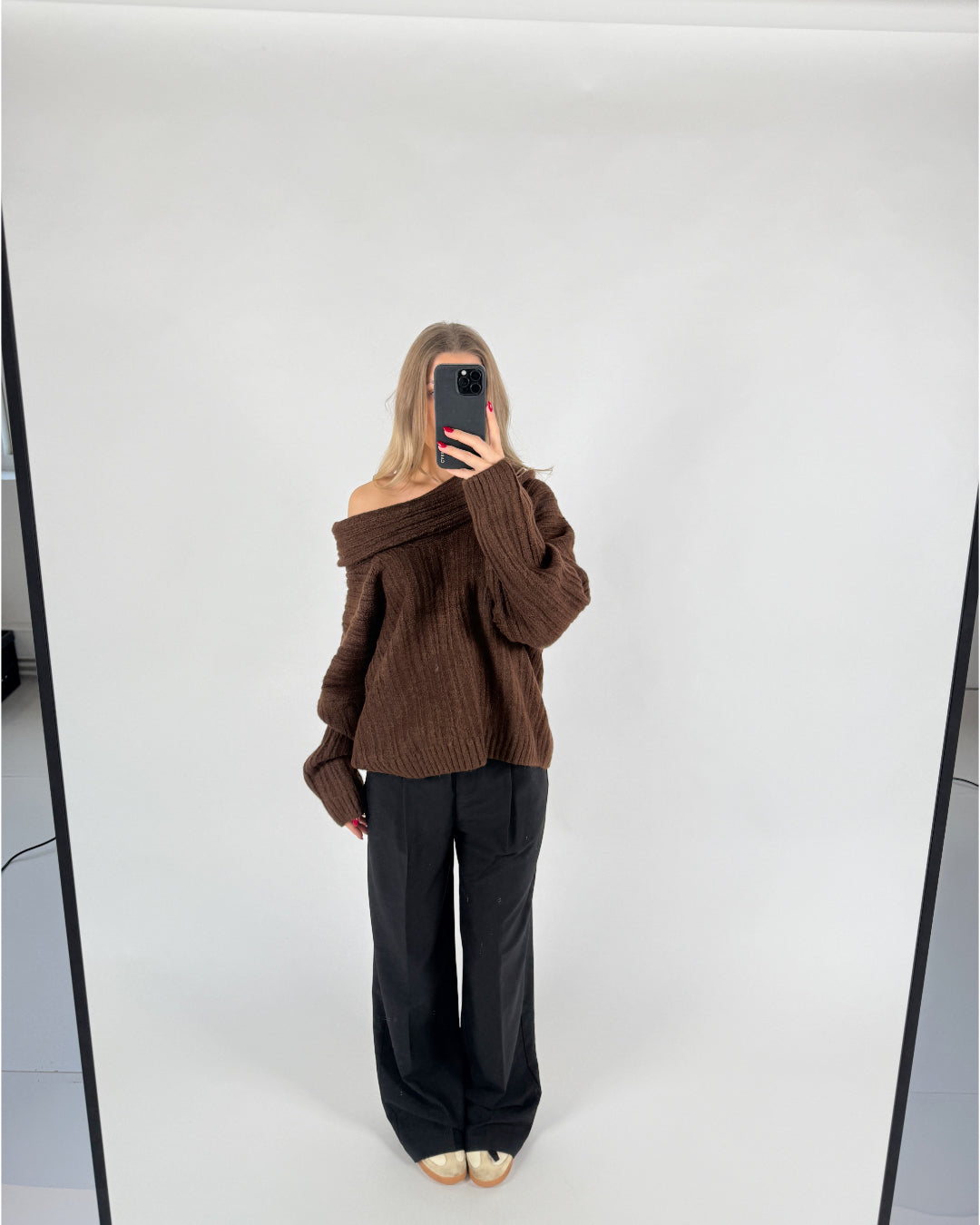 En person iført en brun off-shoulder sweater og sorte bukser tager et spejlbillede. Personen står på en lys baggrund i et minimalistisk rum.