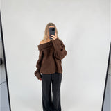 En person iført en brun off-shoulder sweater og sorte bukser tager et spejlbillede. Personen står på en lys baggrund i et minimalistisk rum.
