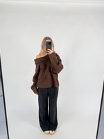 En person iført en brun off-shoulder sweater og sorte bukser tager et spejlbillede. Personen står på en lys baggrund i et minimalistisk rum.