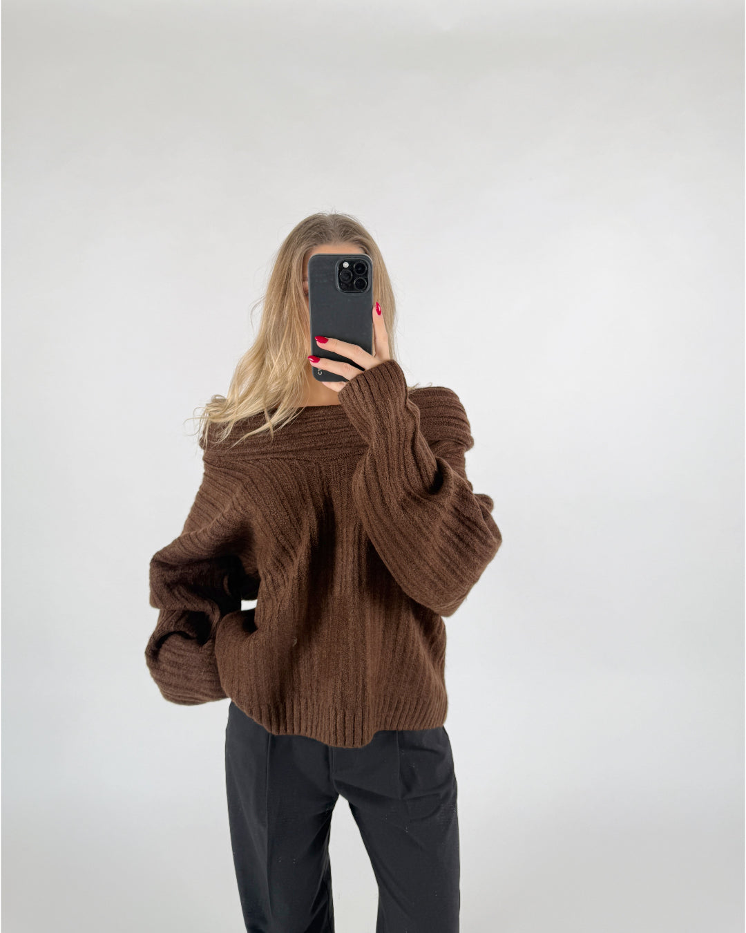 En person iført en brun striktrøje tager et selfie med en sort telefon. Baggrunden er en enkel, lys væg.
