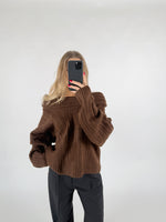 En person iført en brun striktrøje tager et selfie med en sort telefon. Baggrunden er en enkel, lys væg.