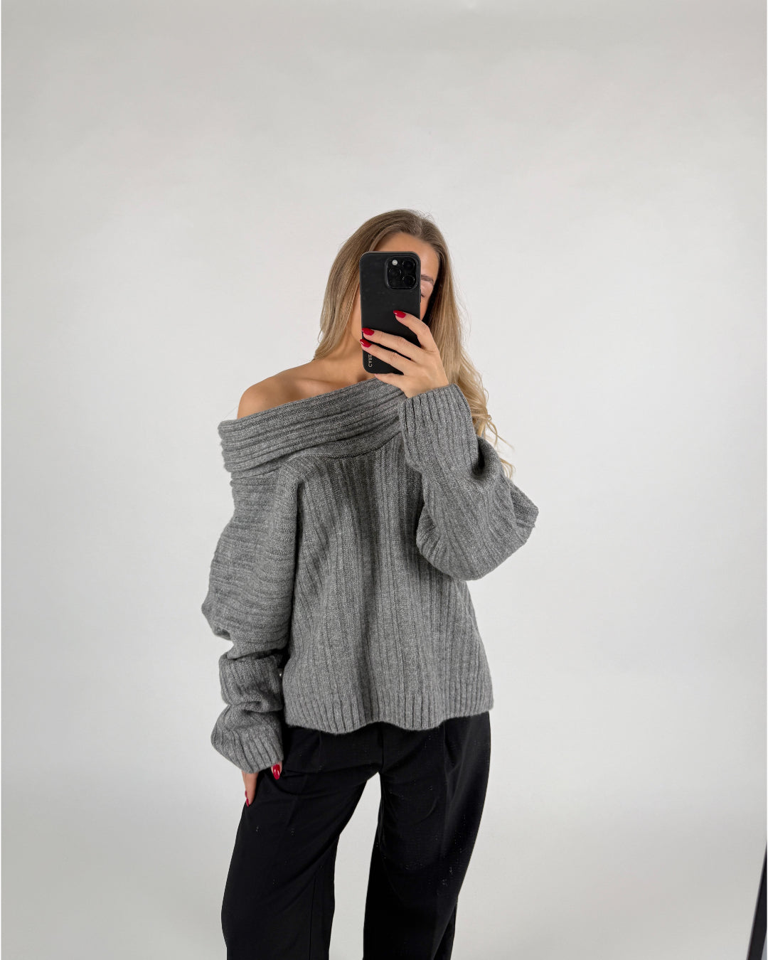 En person iført grå sweater tager et selfie med en smartphone. De står i et neutralt, lys baggrund.