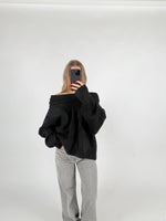 En person iført en sort trøje poserer med en hånd i lommen og en mobiltelefon i den anden, i et minimalistisk, lyst rum.