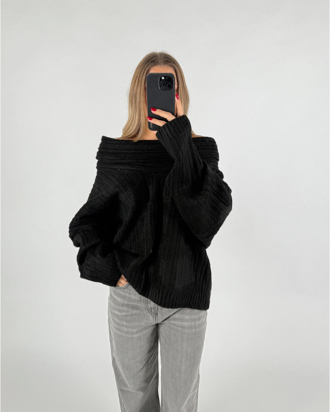 Person tager et spejlselfie iført en sort trøje og grå bukser i et neutralt, lyst rum. Personen har langt hår og rødmalede negle, holder en sort telefon.