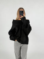 Person tager et spejlselfie iført en sort trøje og grå bukser i et neutralt, lyst rum. Personen har langt hår og rødmalede negle, holder en sort telefon.