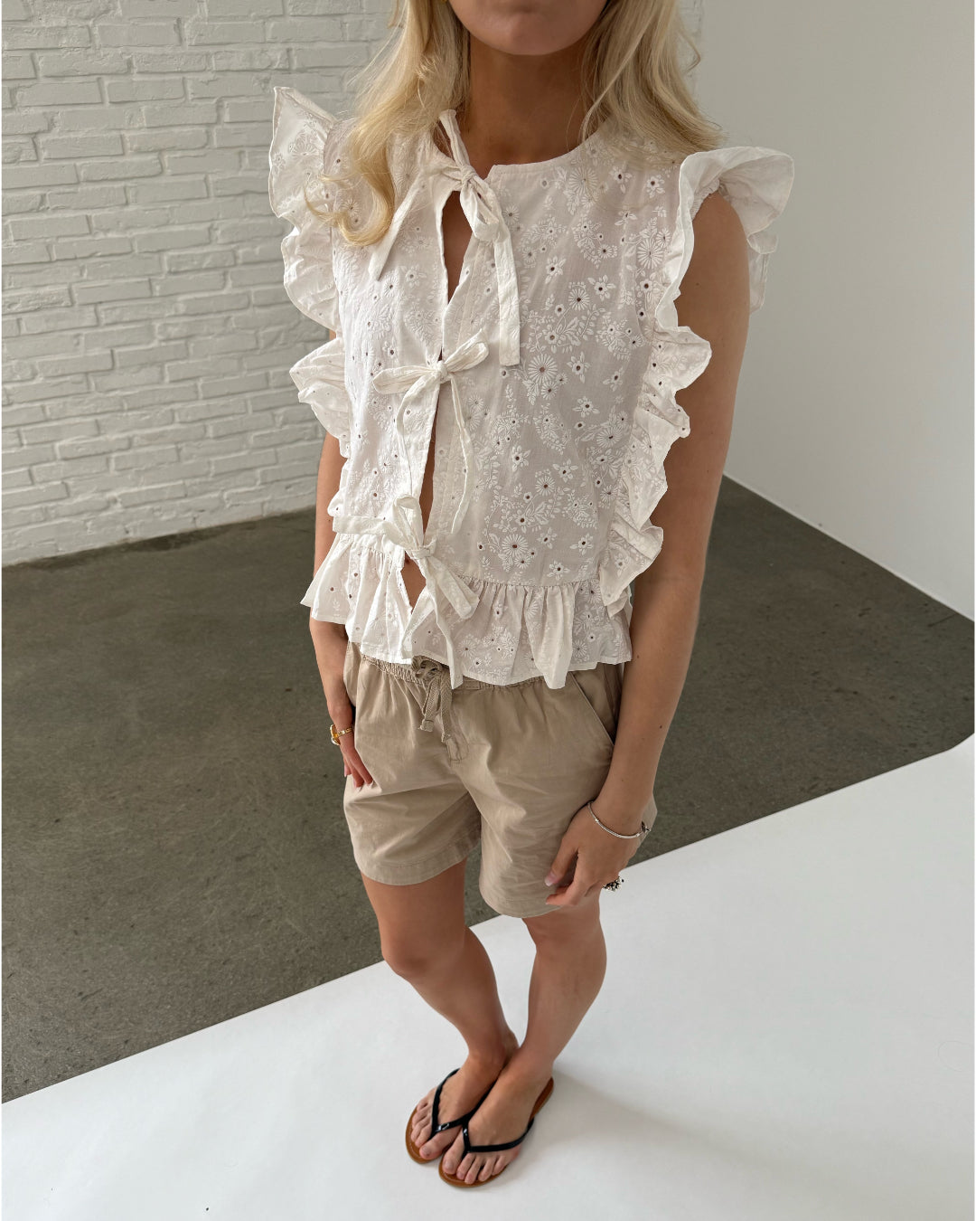 En person iført en hvid, blondebluse og beige shorts står på et betongulv i et rum med hvide vægge. Personen har sorte flip-flops på fødderne.