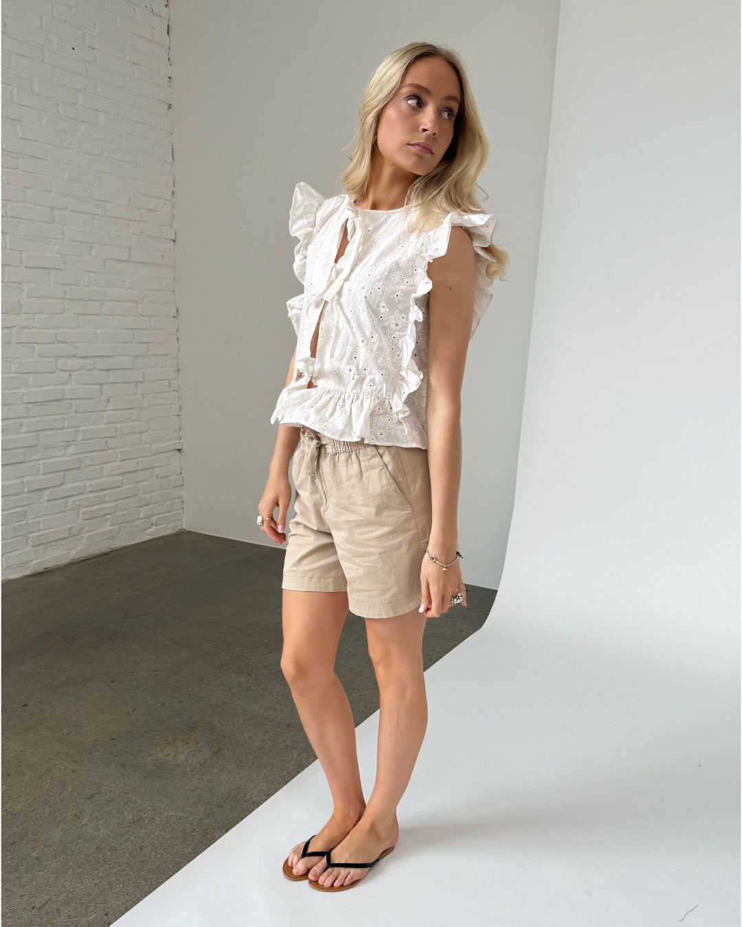 En kvinde iført en hvid, flæsetop og beige shorts står oprejst. Hun kikker til siden i et studie med hvid baggrund og betonvæg.