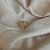 En gylden ring ligger stille på en blød, skinnende lyserød stofoverflade, omgivet af delikate teksturer og lette folder, der fremhæver ringens enkle elegance.