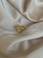 En gylden ring ligger stille på en blød, skinnende lyserød stofoverflade, omgivet af delikate teksturer og lette folder, der fremhæver ringens enkle elegance.