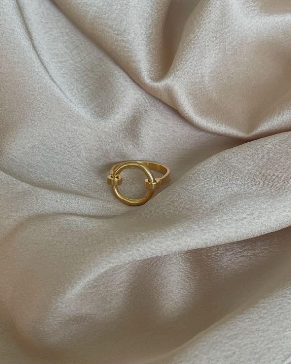 En gylden ring ligger stille på en blød, skinnende lyserød stofoverflade, omgivet af delikate teksturer og lette folder, der fremhæver ringens enkle elegance.