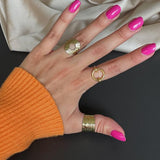 Hånden har tre guldringe og pink neglelak, ligger på en sort overflade ved siden af en beige stofklud. Personen bærer en orange trøje.