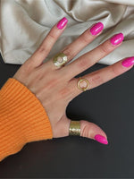 Hånden har tre guldringe og pink neglelak, ligger på en sort overflade ved siden af en beige stofklud. Personen bærer en orange trøje.