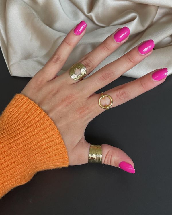 Hånden har tre guldringe og pink neglelak, ligger på en sort overflade ved siden af en beige stofklud. Personen bærer en orange trøje.