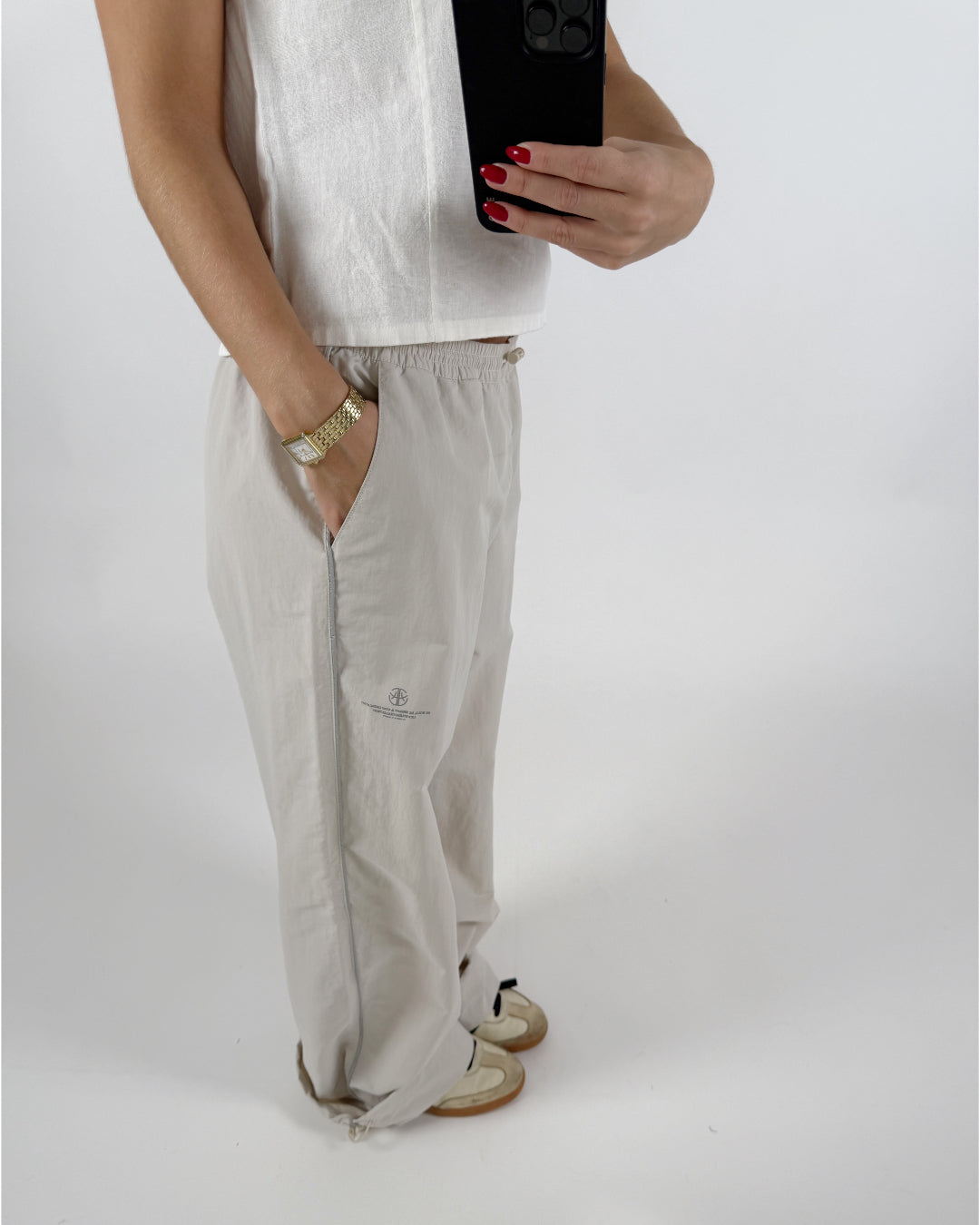Person står i løse, lyse bukser og hvid top, holder en sort telefon med rødneglede fingre, ur på håndled. Kontekst er en lys baggrund. Bukserne har tekst: "Balenciaga Resort 2023."