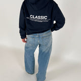 En person iført en mørk hættetrøje med teksten "CLASSIC" og blå jeans står i et lyst rum. Teksten på hættetrøjen fortsætter med mindre skrift under hovedordet.