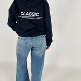 En person iført en mørk hættetrøje med teksten "CLASSIC" og tekst nedenunder står stille. Trøjen bæres over lyseblå jeans i et neutralt rum.