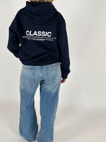 En person iført en mørk hættetrøje med teksten "CLASSIC" og tekst nedenunder står stille. Trøjen bæres over lyseblå jeans i et neutralt rum.