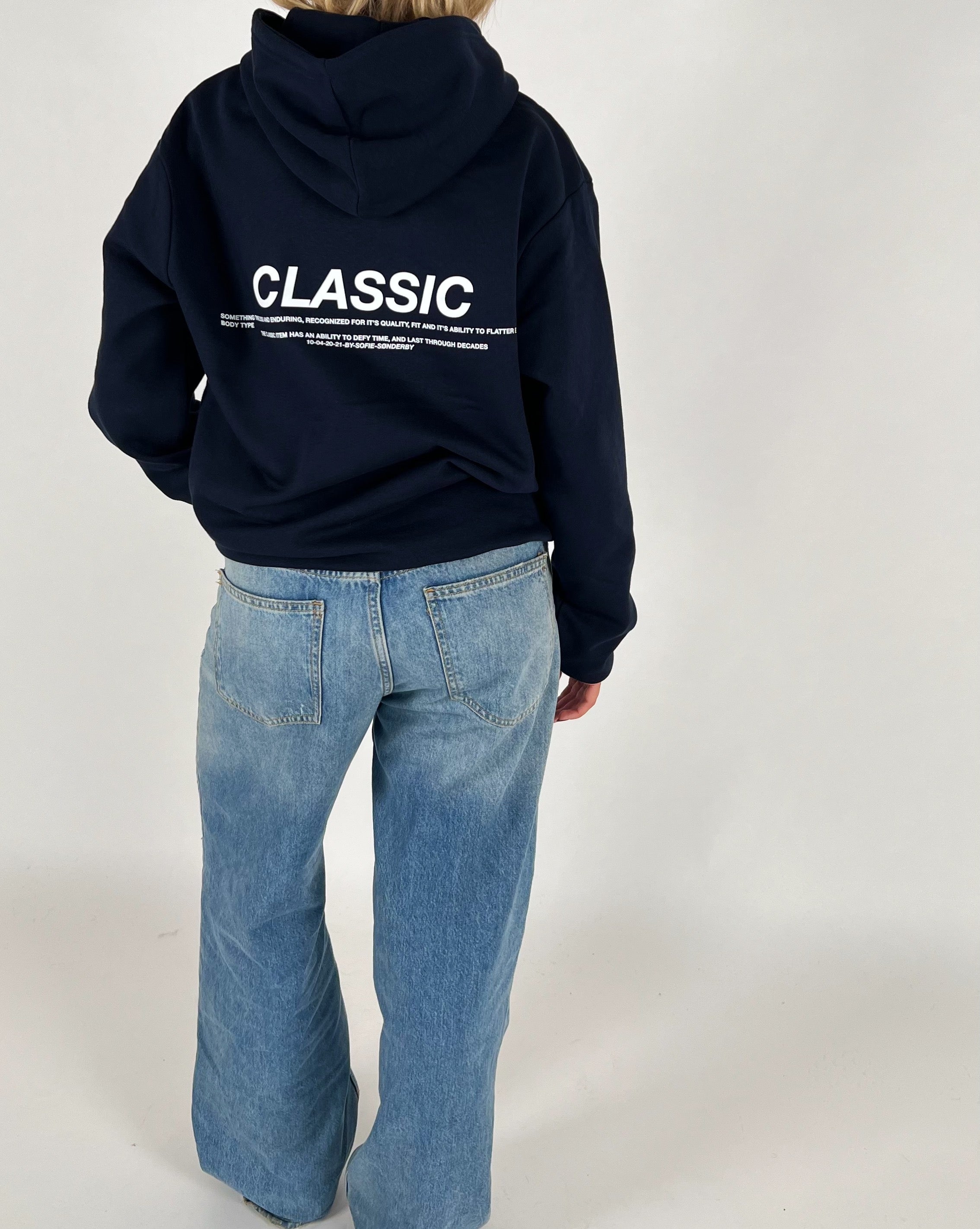 En person iført en mørk hættetrøje med teksten "CLASSIC" og tekst nedenunder står stille. Trøjen bæres over lyseblå jeans i et neutralt rum.