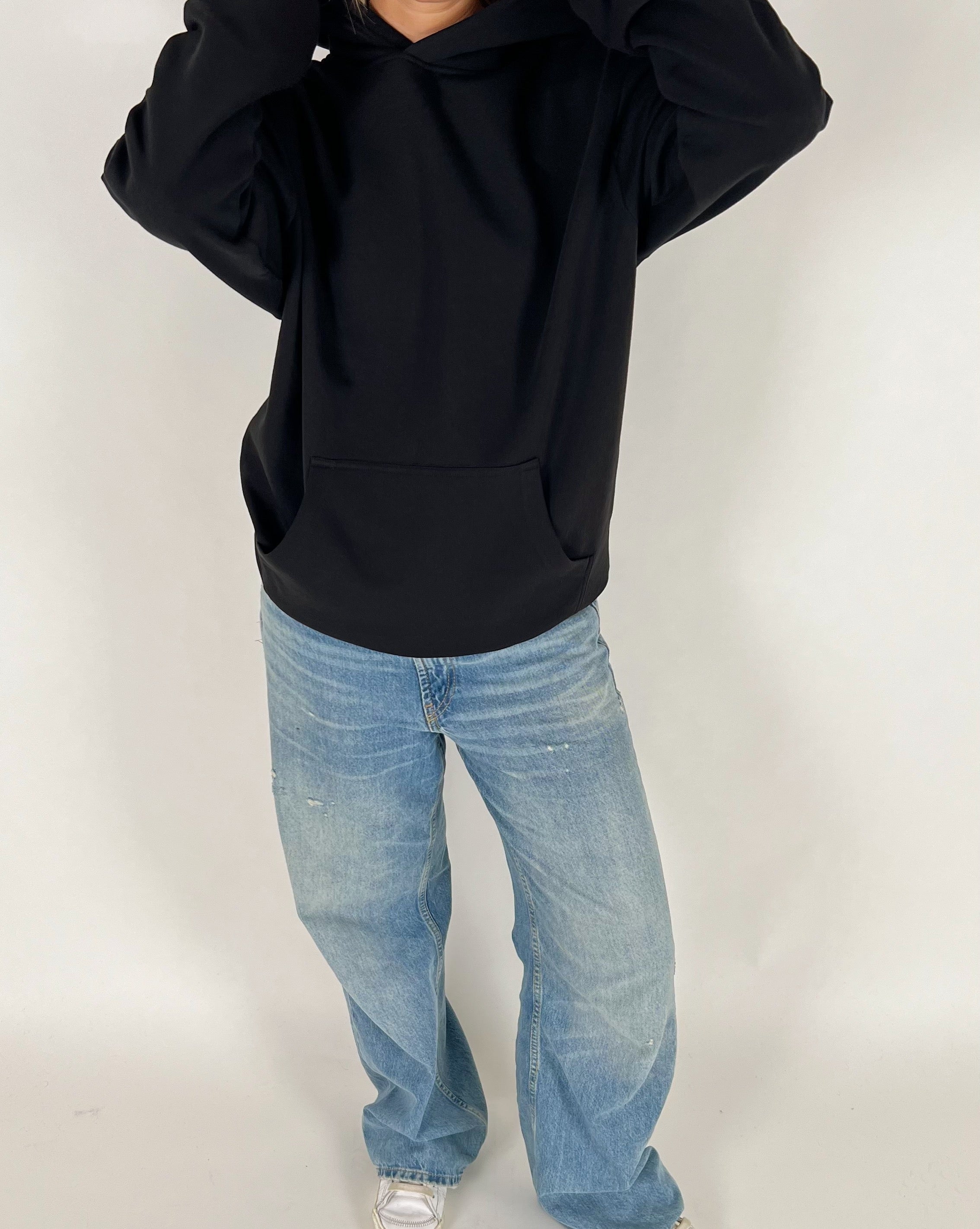En person iført en sort hættetrøje og lyseblå jeans står med hænderne omkring hovedet i et neutralt, indendørs rum.