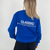En person iført en blå sweatshirt, står med ryggen til i et minimalistisk miljø. Tekst på trøjen: "CLASSIC. Something timeless and enduring, recognized for its quality, fit and its ability to flatter."