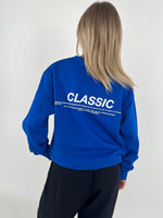 En person iført en blå sweatshirt, står med ryggen til i et minimalistisk miljø. Tekst på trøjen: "CLASSIC. Something timeless and enduring, recognized for its quality, fit and its ability to flatter."