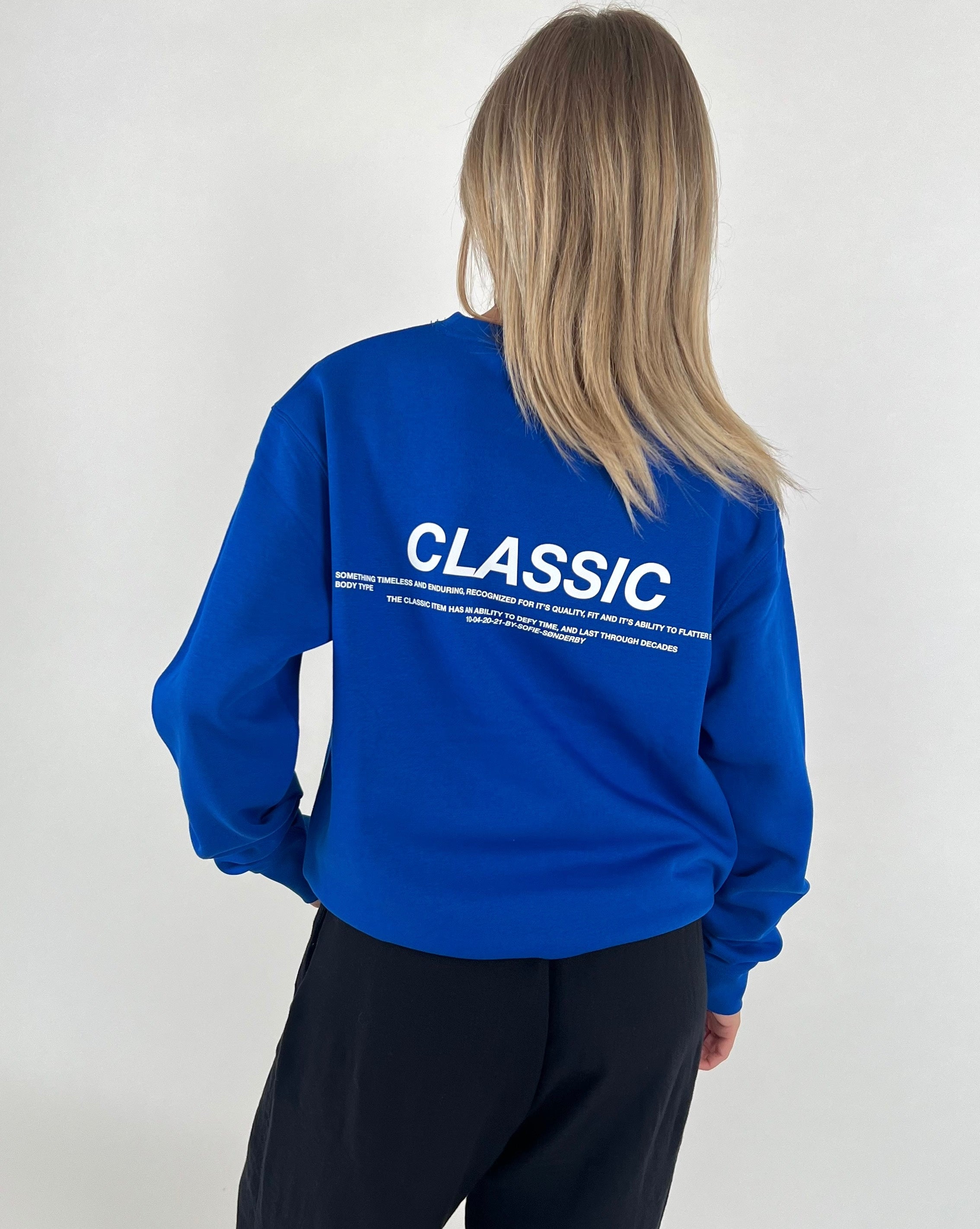 En person iført en blå sweatshirt, står med ryggen til i et minimalistisk miljø. Tekst på trøjen: "CLASSIC. Something timeless and enduring, recognized for its quality, fit and its ability to flatter."