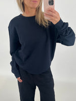 En person iført sort sweatshirt tager et spejlfoto med en smartphone i en lys, minimalistisk baggrund.