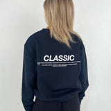 En person står med ryggen til iført en sort sweatshirt med teksten "CLASSIC". Omgivelserne er enkle og neutrale med en lys baggrund.