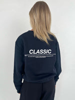En person står med ryggen til iført en sort sweatshirt med teksten "CLASSIC". Omgivelserne er enkle og neutrale med en lys baggrund.