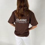 En person står med ryggen til iført en brun t-shirt med teksten "CLASSIC" og yderligere beskrivelse nederst. Personen har langt hår og bærer hvide bukser i et enkelt rum.