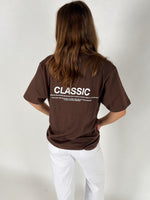 En person står med ryggen til iført en brun t-shirt med teksten "CLASSIC" og yderligere beskrivelse nederst. Personen har langt hår og bærer hvide bukser i et enkelt rum.