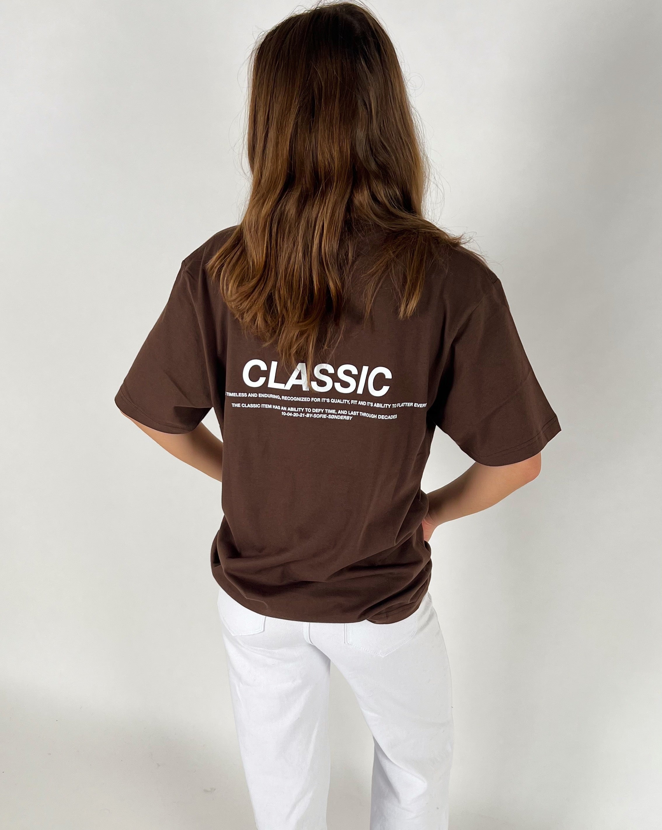 En person står med ryggen til iført en brun t-shirt med teksten "CLASSIC" og yderligere beskrivelse nederst. Personen har langt hår og bærer hvide bukser i et enkelt rum.