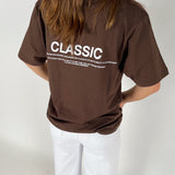 En person iført en brun T-shirt står med ryggen til. På T-shirten står teksten: "CLASSIC. Timeless and enduring, recognized for its quality, fit and its ability to flatter every. The classic item has an ability to defy time, and last through decades. 19-04-20-21-21-Sofie Sonderby." Personen bærer også hvide bukser i et neutralt rum.