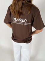 En person iført en brun T-shirt står med ryggen til. På T-shirten står teksten: "CLASSIC. Timeless and enduring, recognized for its quality, fit and its ability to flatter every. The classic item has an ability to defy time, and last through decades. 19-04-20-21-21-Sofie Sonderby." Personen bærer også hvide bukser i et neutralt rum.