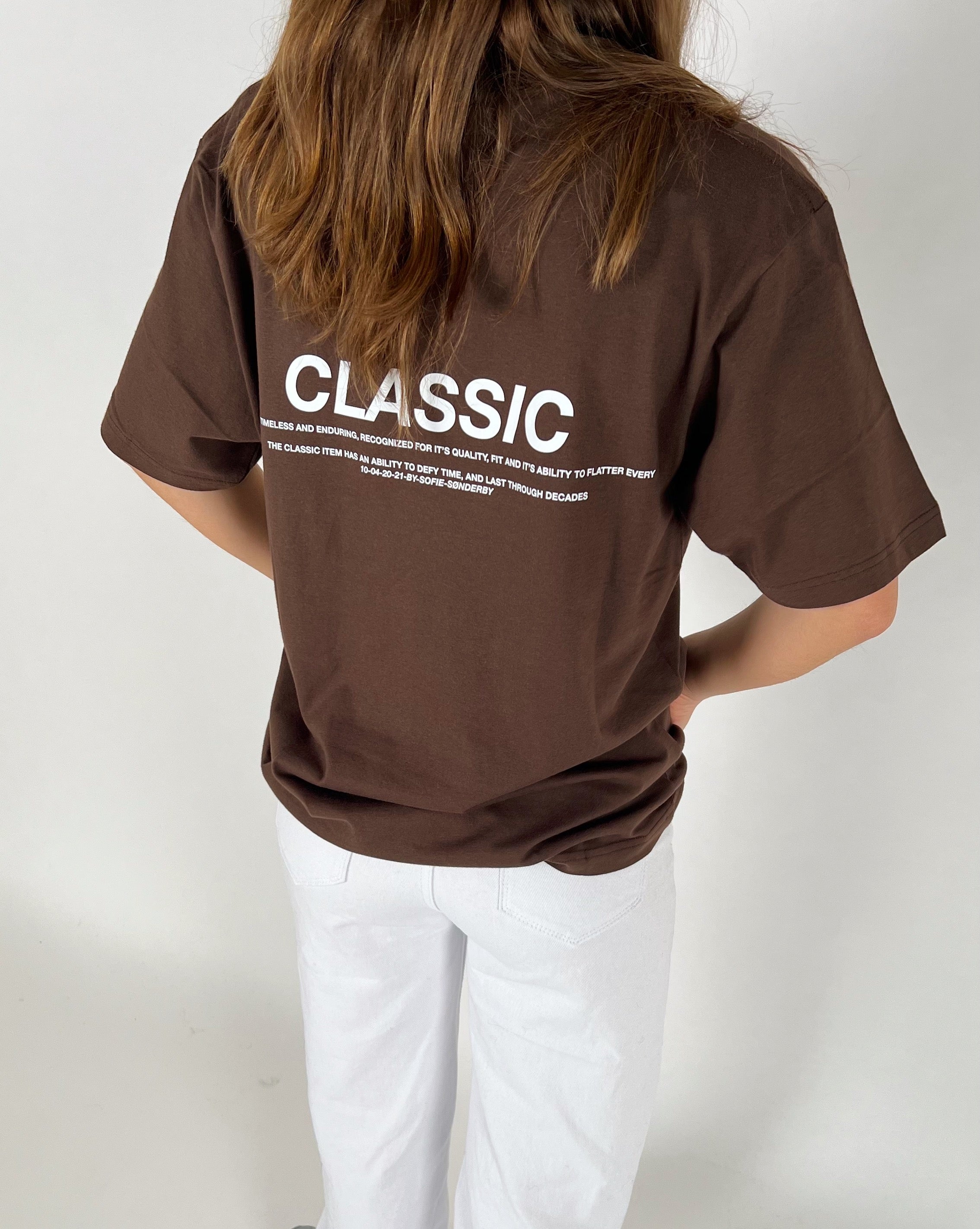 En person iført en brun T-shirt står med ryggen til. På T-shirten står teksten: "CLASSIC. Timeless and enduring, recognized for its quality, fit and its ability to flatter every. The classic item has an ability to defy time, and last through decades. 19-04-20-21-21-Sofie Sonderby." Personen bærer også hvide bukser i et neutralt rum.