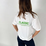 En person står med ryggen til iført en hvid t-shirt med teksten "CLASSIC" i grønt. Personen er i et neutralt rum med hvid baggrund.