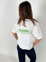En person står med ryggen til iført en hvid t-shirt med teksten "CLASSIC" i grønt. Personen er i et neutralt rum med hvid baggrund.