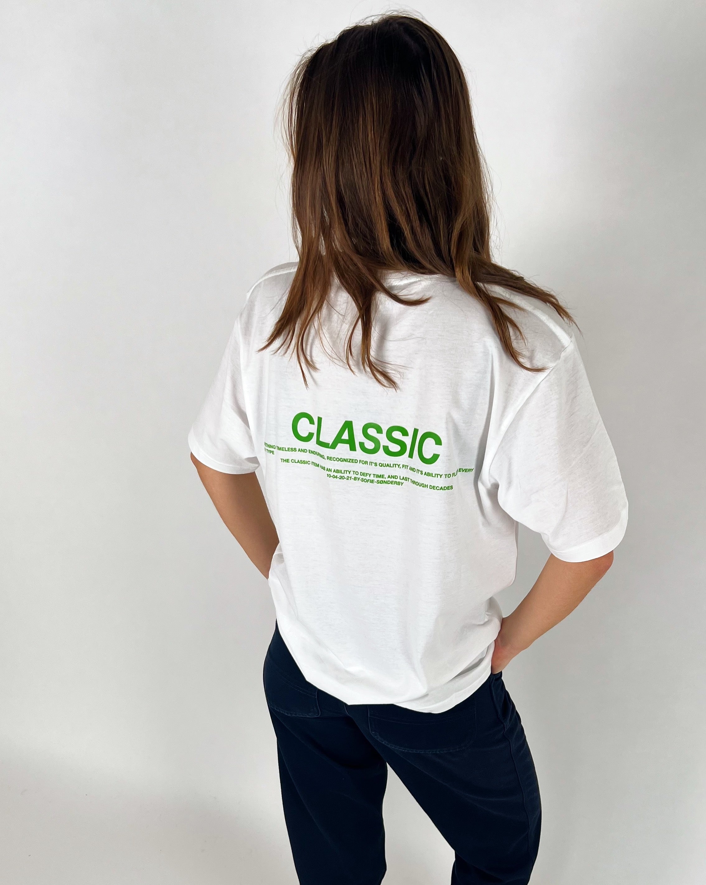 En person står med ryggen til iført en hvid t-shirt med teksten "CLASSIC" i grønt. Personen er i et neutralt rum med hvid baggrund.