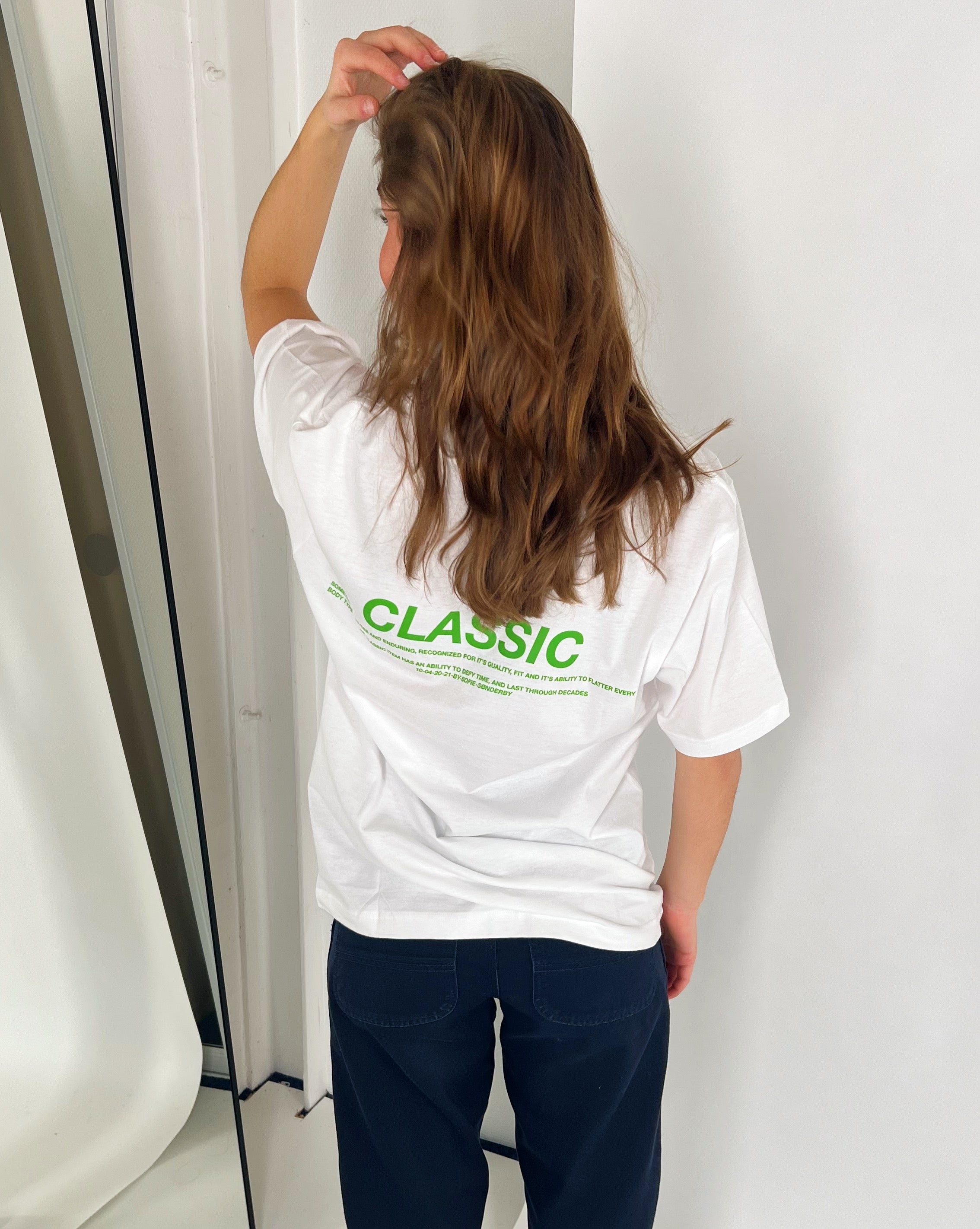 Person i hvid T-shirt står med ryggen til og rører sit hår; teksten på T-shirten siger: “CLASSIC” og noget mere. Personen er i et lyst rum ved en lys væg.