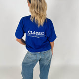 En person står med ryggen til iført en blå T-shirt, hvor der står "CLASSIC" i hvid tekst og løse jeans, mod en neutral baggrund.