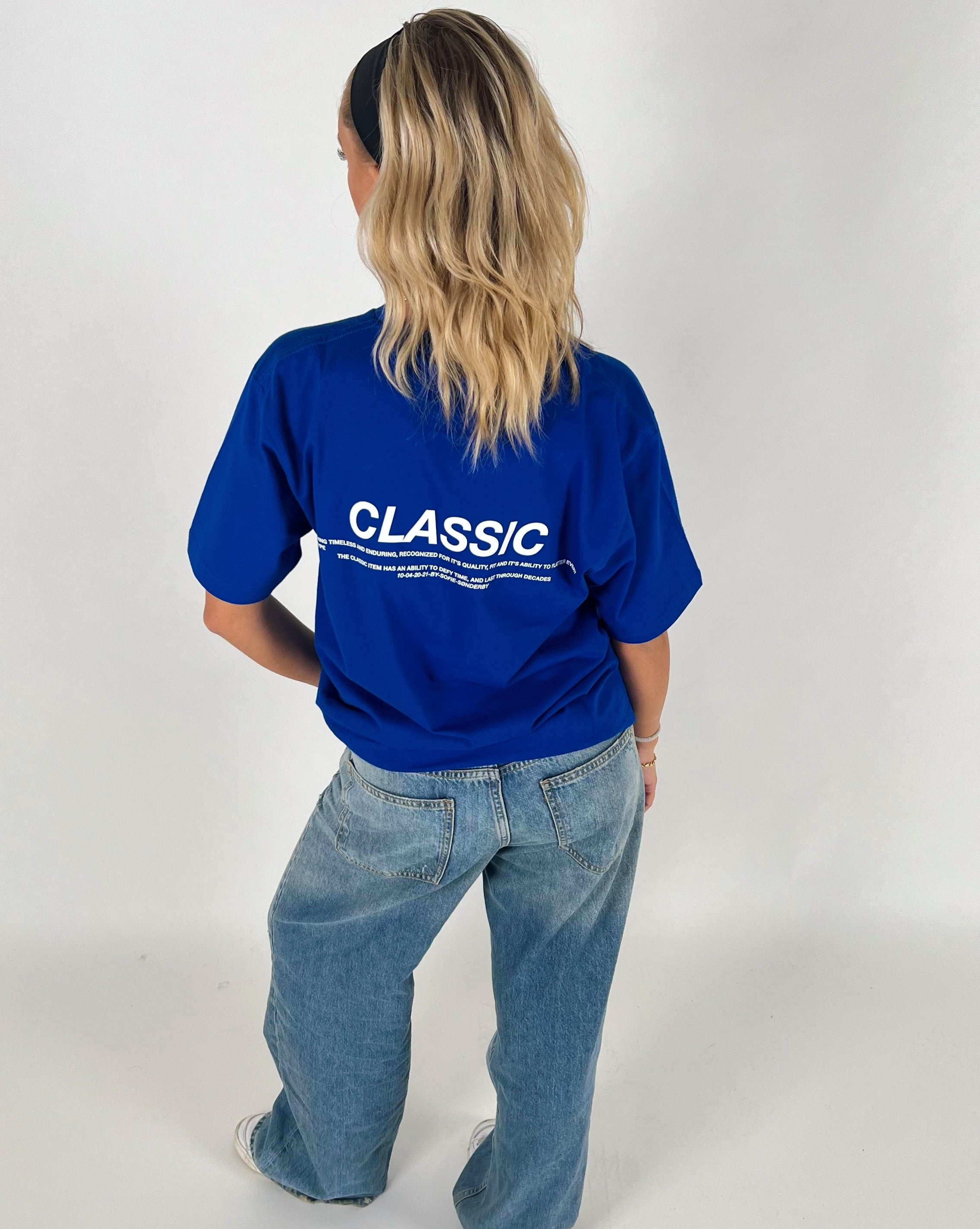 En person står med ryggen til iført en blå T-shirt, hvor der står "CLASSIC" i hvid tekst og løse jeans, mod en neutral baggrund.