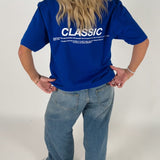 En person iført en blå T-shirt med teksten "CLASSIC" og en længere tekst nedenunder står med hænderne på hofterne i et lyst rum. De har også lyse jeans på.