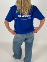 En person iført en blå T-shirt med teksten "CLASSIC" og en længere tekst nedenunder står med hænderne på hofterne i et lyst rum. De har også lyse jeans på.