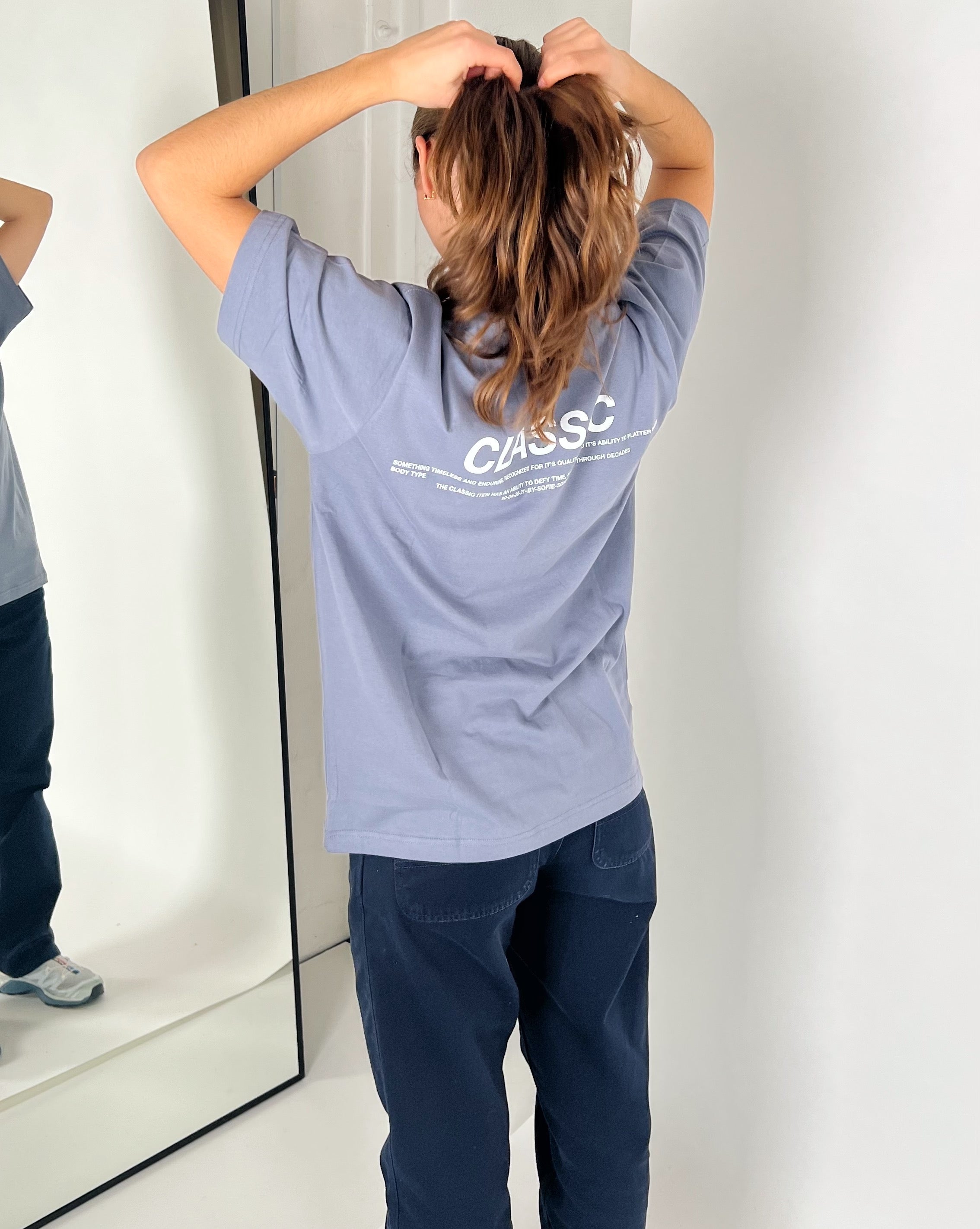 En person i en lyseblå T-shirt arrangerer håret foran et spejl. Spejlingen viser også teksten "CLASSIC" og anden mindre tekst på T-shirtens ryg.